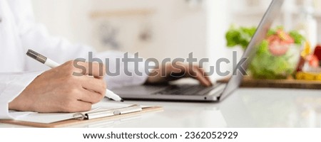Close-up of dietitian hand writing with laptop giving online weight loss and healthy diet consultation with fresh vegetable fruit measuring tape and glass of clean water on desk against Royalty-Free Stock Photo #2362052929