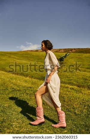 side view of young asian woman in bridal dress and cowboy boots walking on meadow in countryside