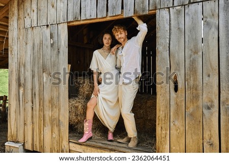 stylish newlyweds, pretty asian bride in cowboy boots and white dress standing with groom in barn