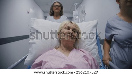 Mature female doctor with young nurse brings senior patient to medical center. Old woman lies on gurney with drip and talks with medics about surgery. Medical staff at work in modern hospital. Royalty-Free Stock Photo #2362107529