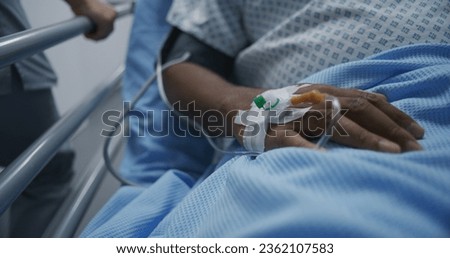 African American patient lies on gurney with catheter on hand. Nurse transports man to hospital room down medical center corridor after surgery. Medical staff at work in modern clinic. Close up. Royalty-Free Stock Photo #2362107583