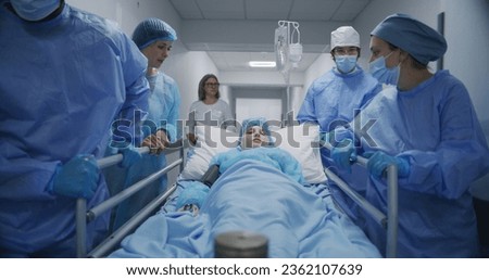 Doctors, paramedics and mom of child push stretcher with sick young girl to operation room. Woman supports lying on gurney daughter. Medical personnel work in emergency department. Tracking shot. Royalty-Free Stock Photo #2362107639