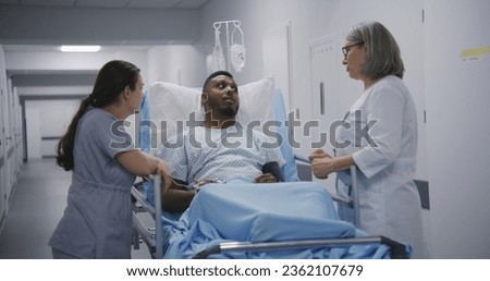Female doctor consults lying on stretcher African American man before operation. Nurse and physician stand near gurney with patient in clinic corridor. Medical personnel work in emergency department. Royalty-Free Stock Photo #2362107679