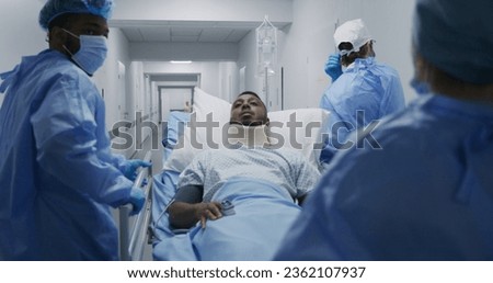 Doctors and paramedics push stretcher with patient to surgery room. African American man with neck brace lies on gurney. Multi ethnic personnel work in clinic. Slow motion. Back view. Tracking shot. Royalty-Free Stock Photo #2362107937