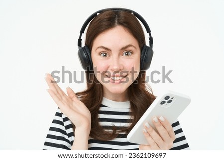 Portrait of beautiful young woman, wearing headphones, holding smartphone, smiling and looking happy, isolated on white background.