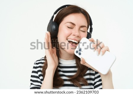 Portrait of happy girl playing karaoke app, singing into smartphone microphone, wearing wireless headphones, white background.