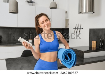 Image of strong and fit fitness instructor, woman standing at home with rubber mat and smartphone in hands, has an app for workout indoors.