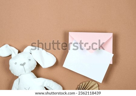 Pink envelope with empty white card for text and white plush bunny on beige background. Blank invitation or greeting card mockup with copy space