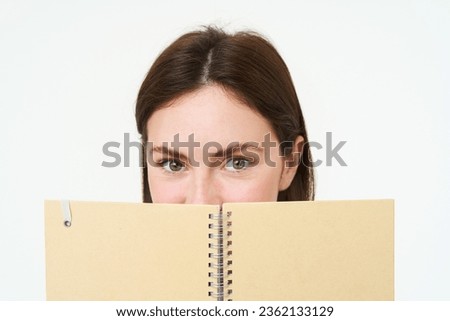 Young woman holds notebook daily planner next to her face, writing down homework, making notes, looking thoughtful, standing over white background.