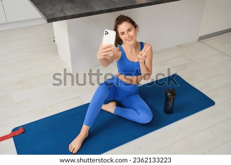 Portrait of stylish, smiling young brunette woman, wears blue sportswear, poses on yoga mat with smartphone, shows peace sign and takes selfie, does workout at home.