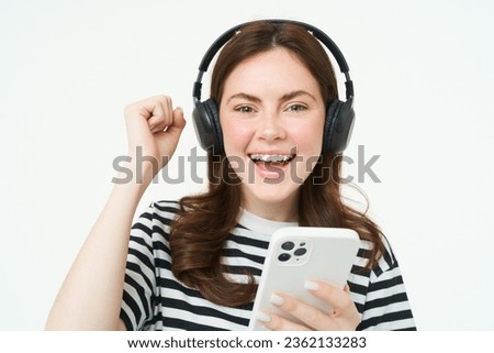 Enthusiastic girl in headphones, holding smartphone, smiling and cheering, celebrating, raising hand up, achieve goal, standing over white background.