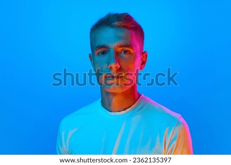 Portrait of young good-natured, calm man, student in white t-shirt looking at camera isolated on blue background in red neon light. Concept of emotions, facial expression, youth, aspiration, sales. Royalty-Free Stock Photo #2362135397