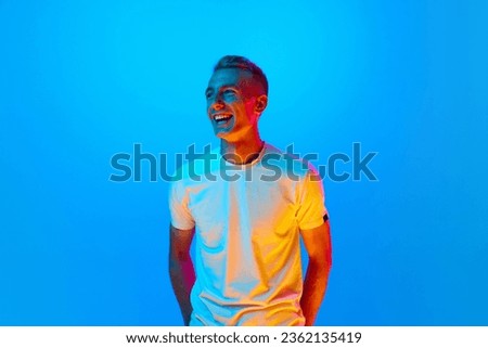 Portrait of young happy, smiling, laughing man, student in white t-shirt looking at camera isolated on blue background in red neon light, filter. Concept of beauty, art, fashion, youth, sales and ad.
