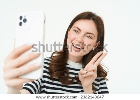 Portrait of young woman taking selfies on smartphone, posing for photo,. using mobile phone app for taking funny and cute pictures, isolated on white background.