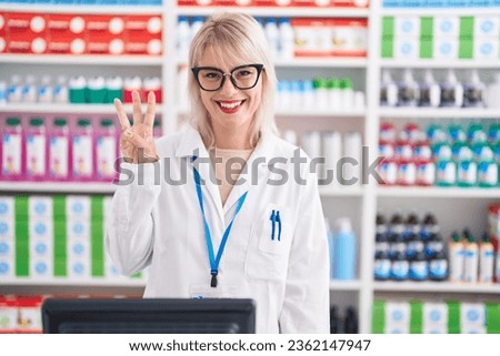 Young caucasian woman working at pharmacy drugstore showing and pointing up with fingers number three while smiling confident and happy. 