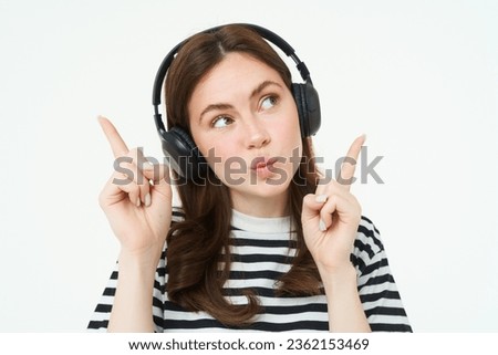 Portrait of young brunette woman in headphones, listening music in headphones, pointing fingers up, showing banner, advertisement, isolated over white background.