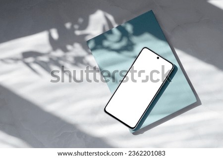 Mobile telephone empty display mock up, blue notebook on white marble table with natural abstract sun light shadows background. Aesthetic business brand template. Royalty-Free Stock Photo #2362201083