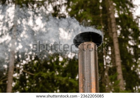 Against the backdrop of a fuzzy forest, smoke rises from a metal chimney, giving the peaceful landscape an atmospheric feel
