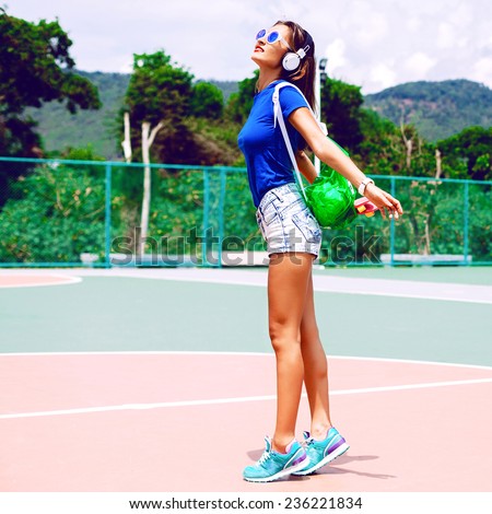 Outdoor lifestyle fashion image of stylish fit woman posing on sports ground, wearing stylish summer clothes sunglasses and neon backpack, listening music in earphones. amazing view on mountains.