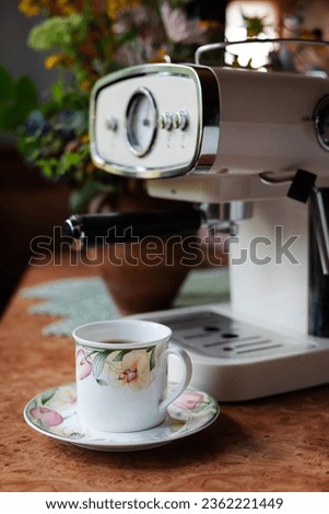 A cup of coffee on the table, a coffee maker in the background and a bouquet of flowers in a vase.