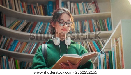 Pretty caucasian young stylish female student in glasses reading textbook while learning in library and flipping pages. Beautiful woman searching for information in book. Literature concept.