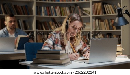 Beautiful blonde female in headset talking, writing and videochatting on laptop in library. Woman speaking, taking notes and having videochat online via webcam. Video call. Noting information.