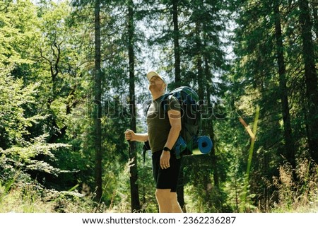 Adult man with backpack and hiking stick walking in green forest in daylight
