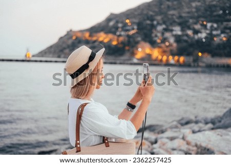 Young woman in a straw hat taking a picture with her smartphone. Sea, evening lights, hills on the background 