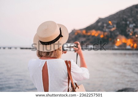 Woman in white dress and straw hat taking a picture from the back. Sea, evening lights. hills on the background 