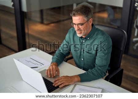 Smiling busy middle aged professional business man working on laptop at office desk. Older mature Indian happy male entrepreneur worker typing on computer, executive manager using pc in office.