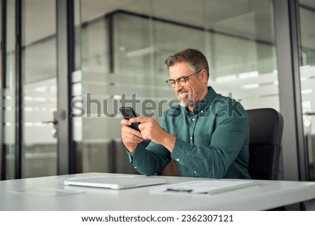 Smiling mid aged businessman executive using cell phone at work desk. Happy busy mature older professional business man manager investor checking finance apps on smartphone in office looking at mobile