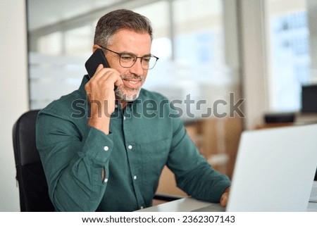 Middle aged businessman making call on cellphone using laptop in office. Smiling busy mature old professional business man manager talking on phone working at laptop computer communicating at work. Royalty-Free Stock Photo #2362307143