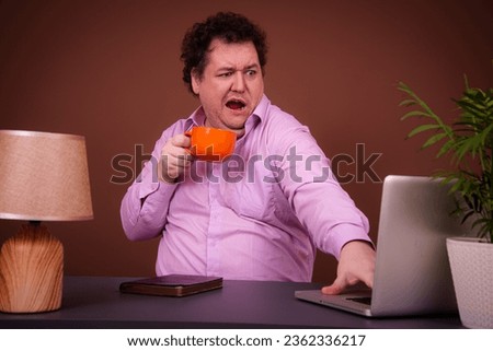 Funny fat man posing at a table with a laptop. Hard work online early morning Brown background.