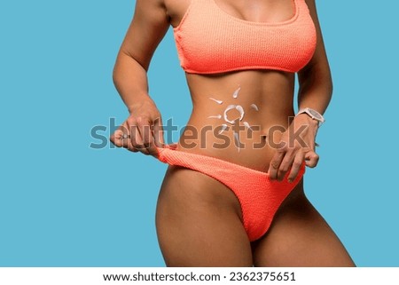 Woman with moisturizing sunscreen cream on tanned belly in form of sun. Slender girl in orange swimwear on blue background. Perfect slim toned young body female