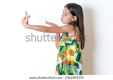 Beautiful kid girl standing over blue background blows air kiss at camera of smartphone and takes selfie, sends mwah via online call.