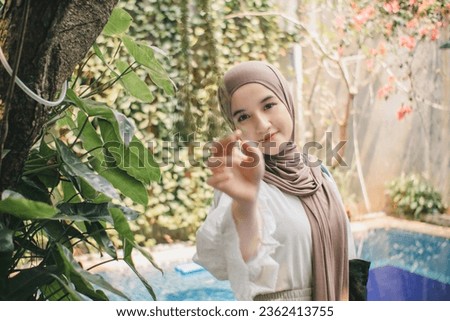 A woman wearing a beautiful hijab smiled sweetly with her head slightly tilted and her hands holding a branch above the cool swimming pool