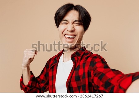 Close up young man of Asian ethnicity he wear red shirt casual clothes doing selfie shot pov on mobile cell phone do winner gesture isolated on plain pastel light beige background. Lifestyle concept