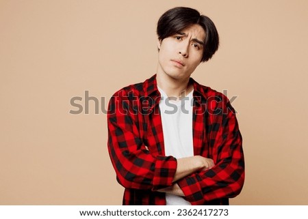 Young frowning upset frustrated sad man of Asian ethnicity wear red checkered shirt casual clothes hold hands crossed folded isolated on plain pastel light beige background studio. Lifestyle concept
