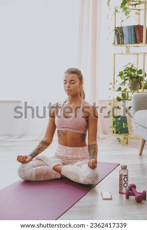 Beautiful attractive young strong sporty fitness woman wearing pink tracksuit doing yoga exercises sitting relaxing meditating trying to calm down keeping eyes closed stretching on mat at home indoor