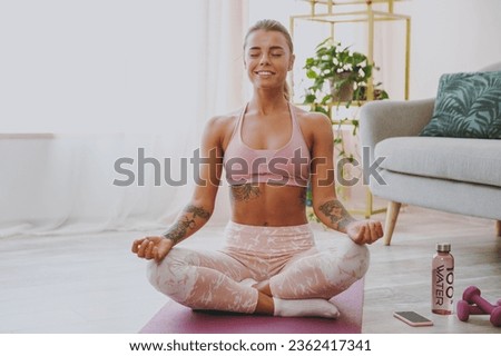 Smiling attractive young strong sporty fitness woman wearing pink tracksuit doing yoga exercises sitting relaxing meditating trying to calm down keeping eyes closed stretching on mat at home indoor