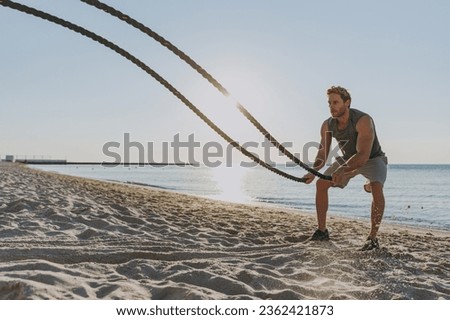 Young strong sporty athletic toned fit sportsman man in sports clothes warm up train work out with battle ropes at sunrise sun dawn over sea beach outdoor on sand beach seaside in summer day morning Royalty-Free Stock Photo #2362421873
