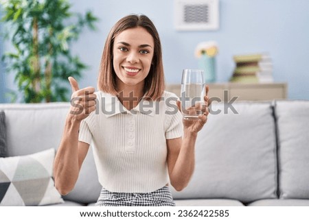 Hispanic woman drinking glass of water smiling happy and positive, thumb up doing excellent and approval sign 