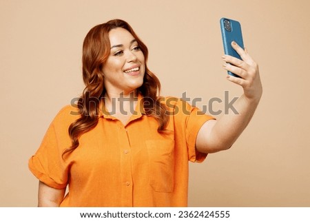 Young ginger chubby overweight woman wear orange shirt casual clothes do selfie shot on mobile cell phone post photo on social network isolated on plain pastel light beige background studio portrait