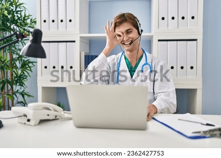 Young doctor man working on online appointment smiling happy doing ok sign with hand on eye looking through fingers 
