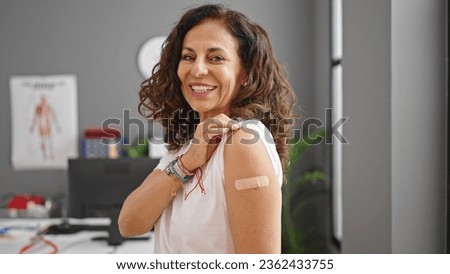 Middle age hispanic woman smiling confident standing with band aid on arm at clinic