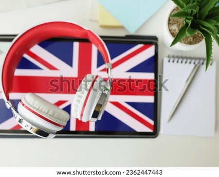 Tablet with image of British flag with headphones and notebook with pen lie on table. Learn British English remotely via app concept
