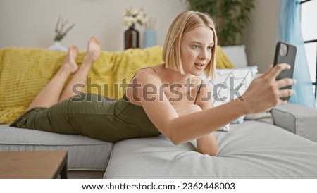 Young blonde woman taking selfie picture with smartphone lying on the sofa at home