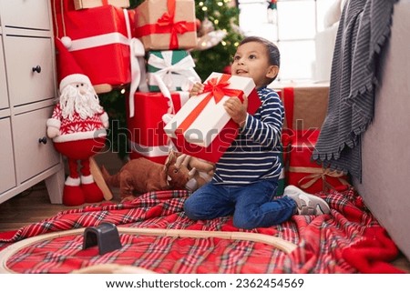 Adorable hispanic toddler holding gift sitting on floor by christmas tree at home