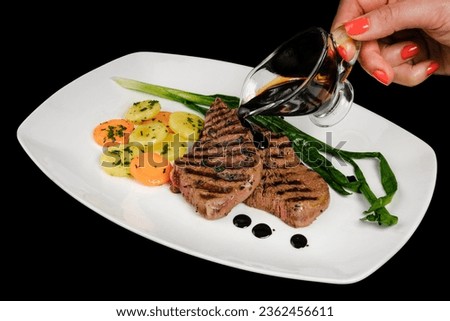 Pouring sauce over juicy piece of meat. The chef's hand decorating the food pours sauce over the steak. A pieces of steak with grilled vegetables. Isolated on black