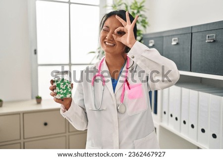 Young asian doctor woman holding pills smiling happy doing ok sign with hand on eye looking through fingers 